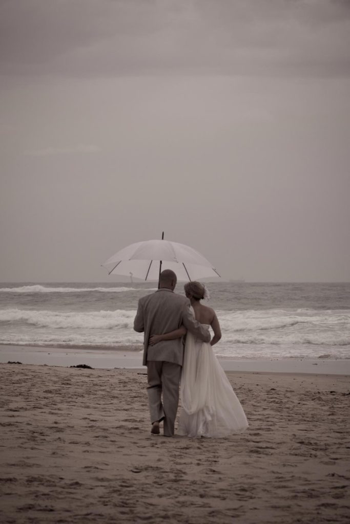bride and groom walk on the beach with umbrella